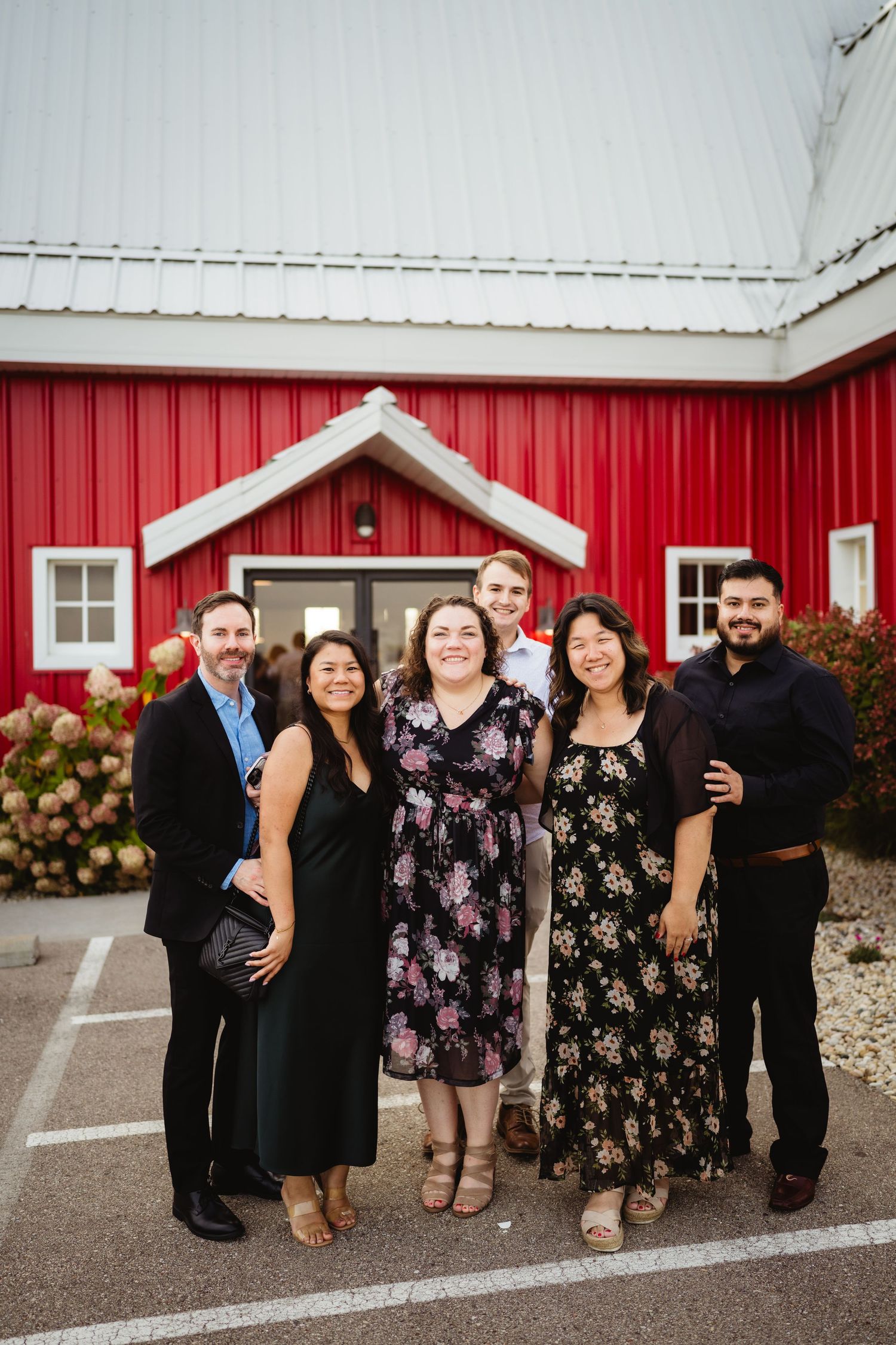 Wedding guests pose for group photos outside a charming red barn on a cloudy day.