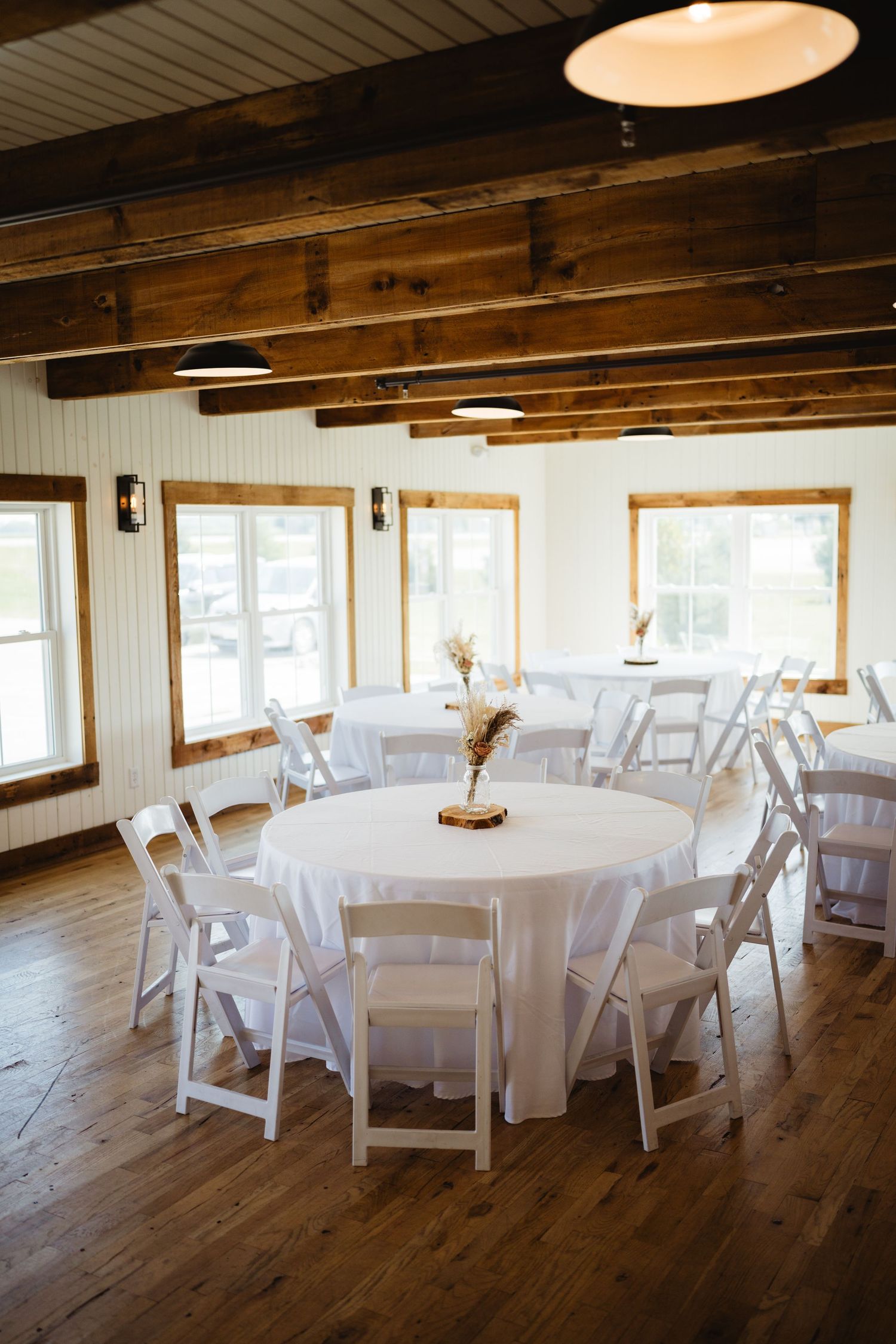Rustic event venue interior with exposed wooden beams, white round tables, chairs and natural lighting from windows.