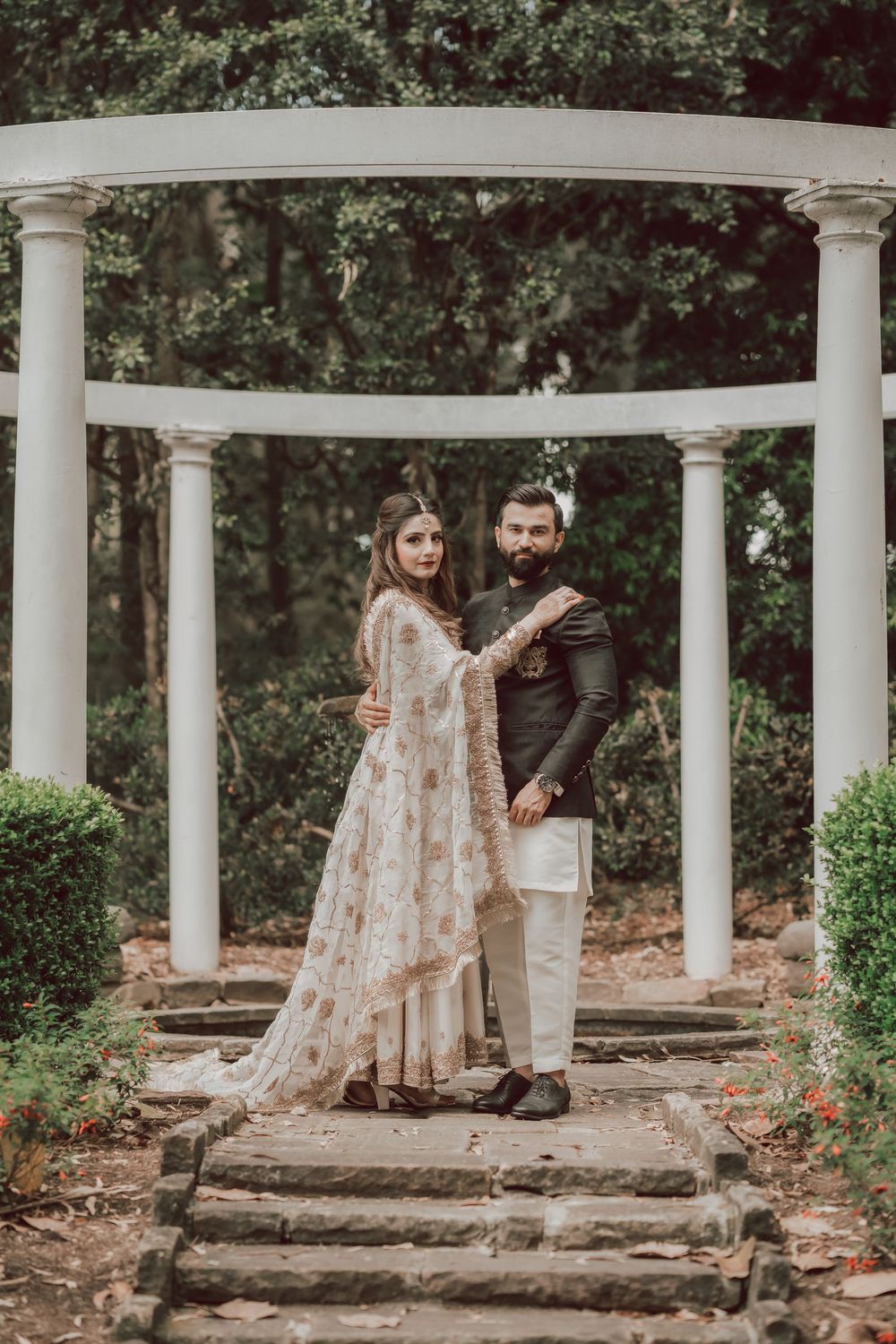 A romantic couple poses together in a serene garden setting with white columns and natural greenery in the background.