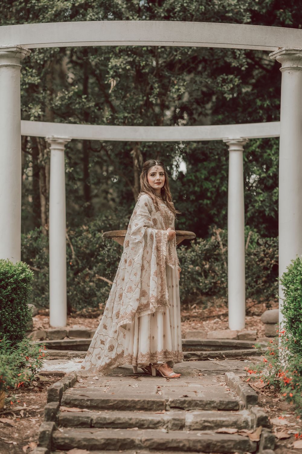 A long floral dress flows elegantly between white columns in a romantic outdoor garden setting.