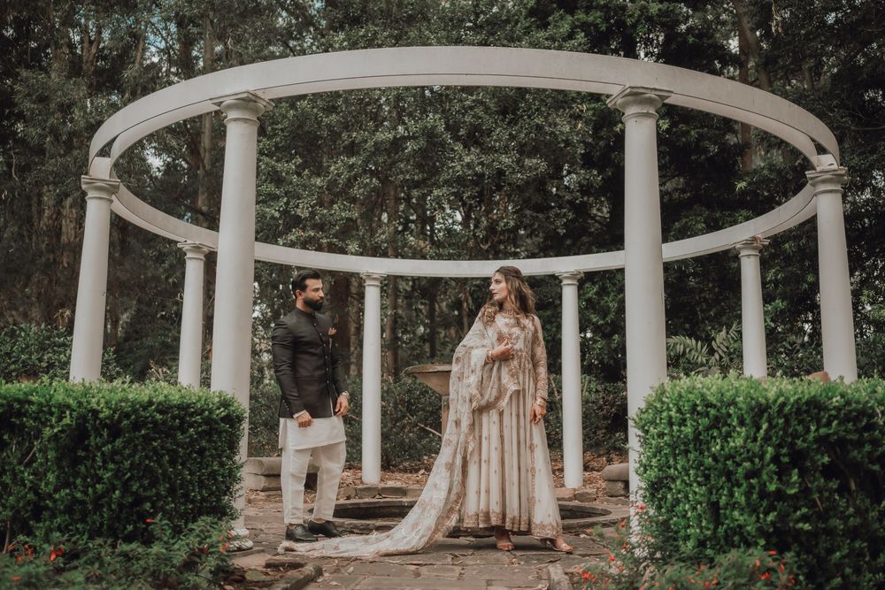 Elegant couple poses under a white columned gazebo surrounded by lush greenery during a romantic outdoor photoshoot.