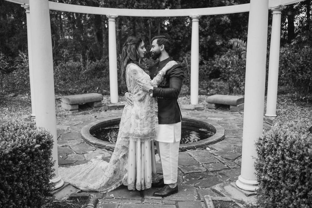 Two people stand in a columned garden gazebo surrounded by landscaping on a sunny day.