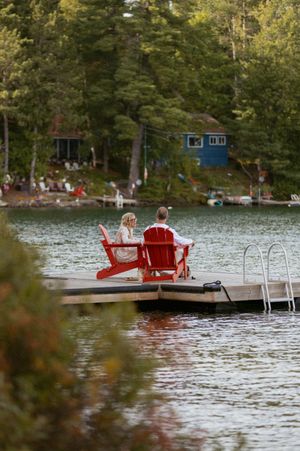 Red Adirondack chairs sit on a wooden dock overlooking a peaceful lake surrounded by trees.