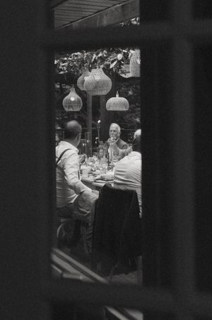 Dimly lit dinner party scene viewed through window panes with hanging pendant lights.
