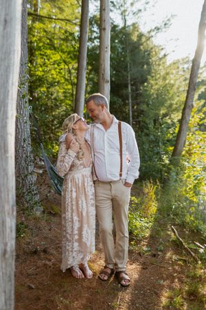 A romantic couple in vintage-inspired attire share a tender moment on a wooded path during golden hour.