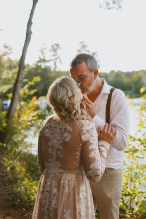 A couple embraces in lace and suspenders against a sunlit forest backdrop near a lake.