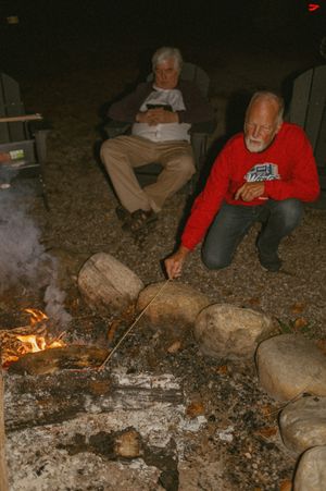 Two people sit by a rustic campfire surrounded by stones in the evening darkness.