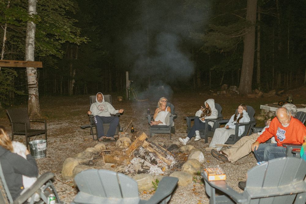 A group gathers around a large campfire in camping chairs during nighttime in the woods.