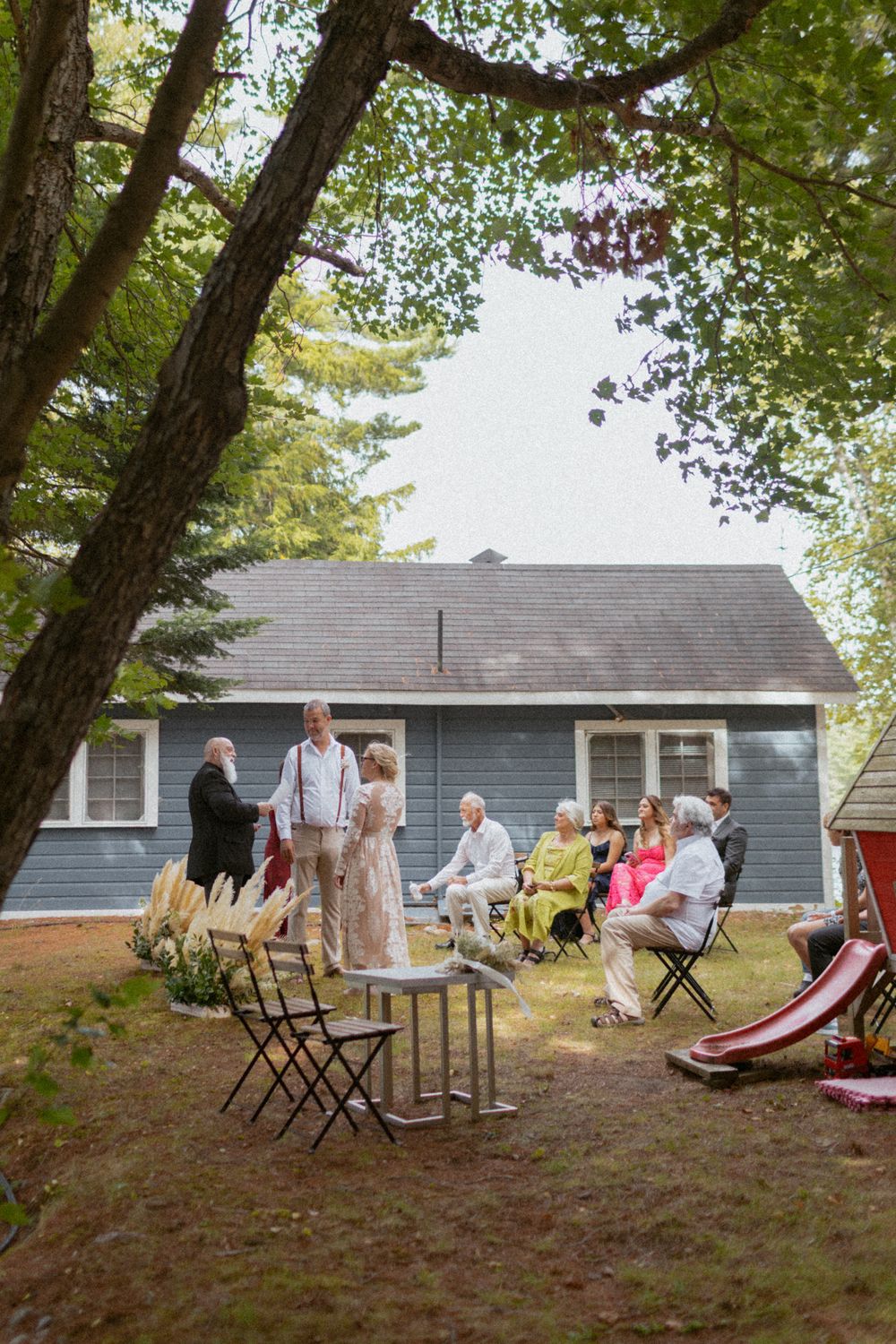 Outdoor gathering with chairs set up in front of a gray house surrounded by trees.