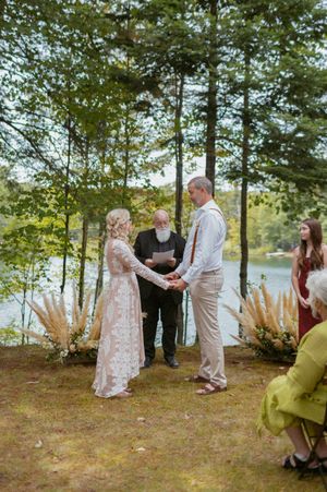 Intimate wedding ceremony taking place under tall trees by a lakeside.