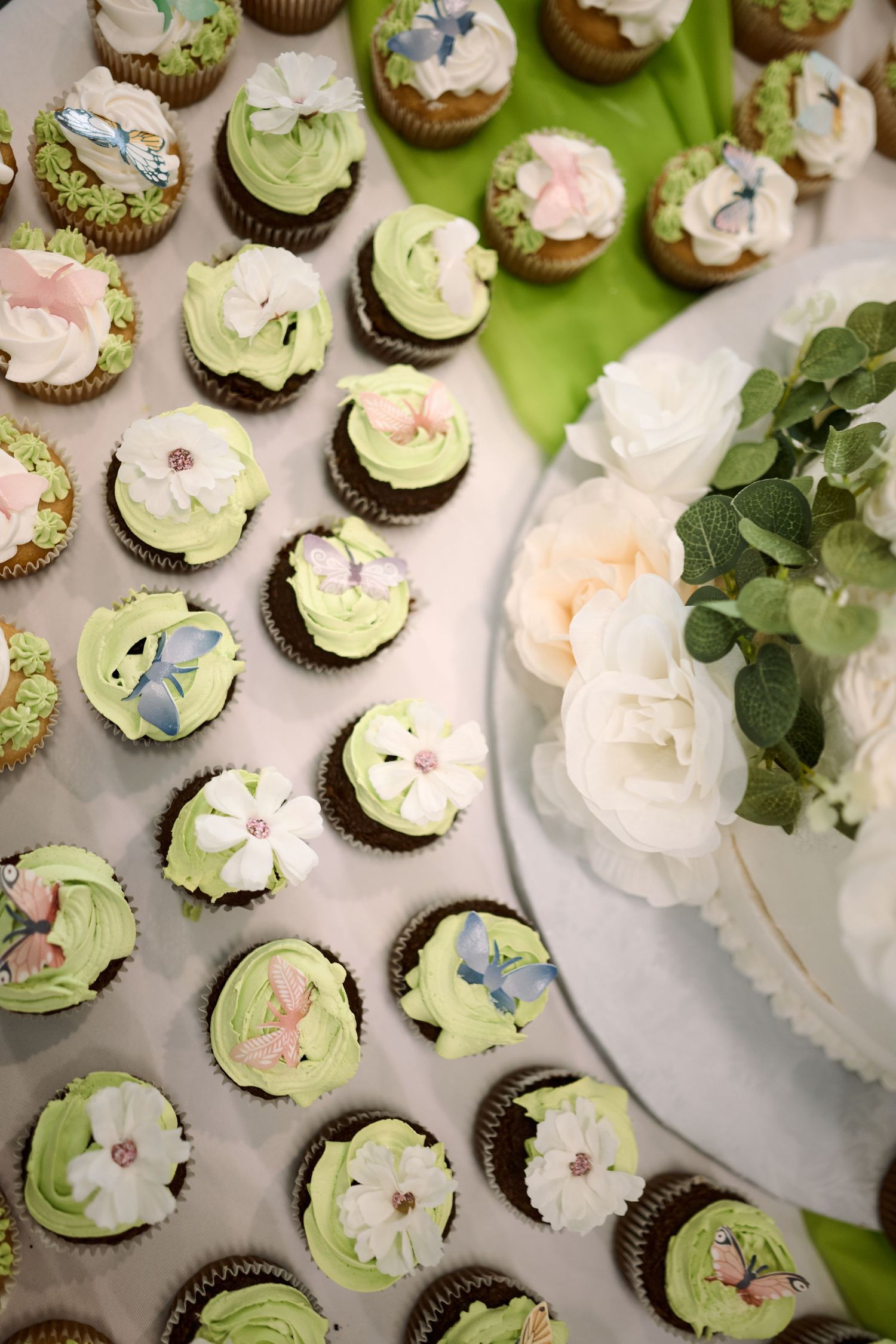 Elegant mint green cupcakes decorated with butterflies and flowers arranged beside white roses and greenery on a table.