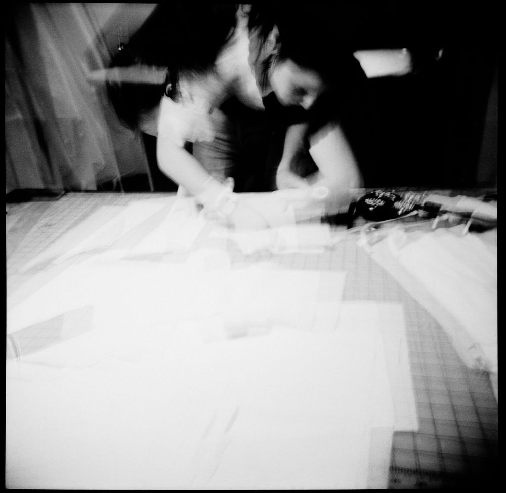 Two women work together cutting white fabric on a large table in a black and white motion-blurred photo.