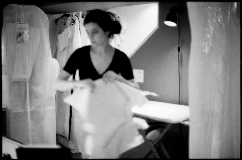 A woman in a black top handles white fabric near garment bags in a dimly lit black and white workshop.