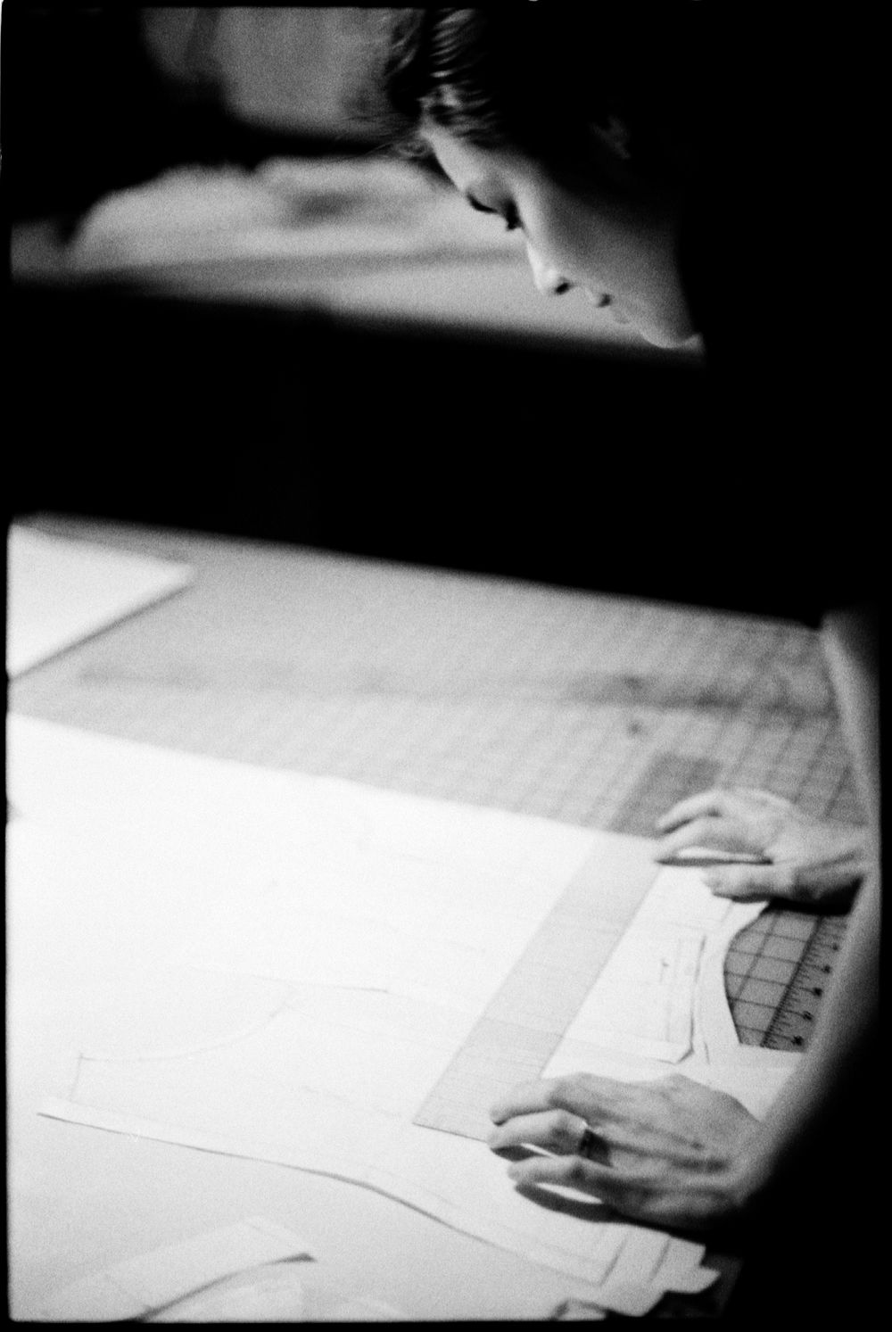 A fashion designer traces sewing patterns on paper using a ruler on a large work table in black and white.