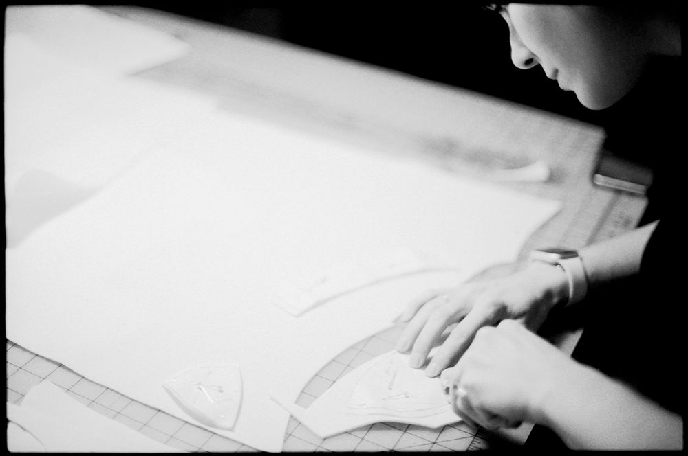 A designer traces curved sewing pattern pieces on paper at a drafting table in a black and white photo.