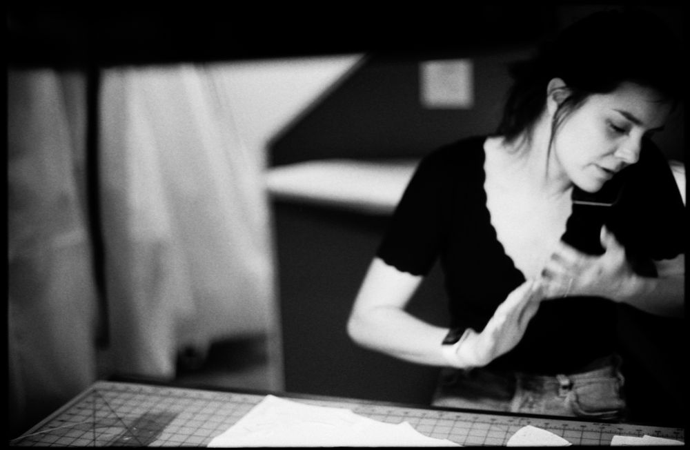 A woman in a black top works with white fabric at a cutting table in a black and white workshop photo.