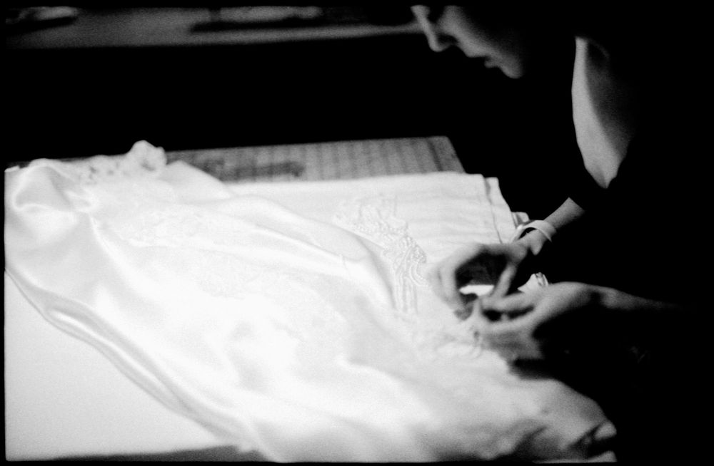 A dressmaker carefully marks or cuts intricate white lace fabric on a dark work surface in black and white.