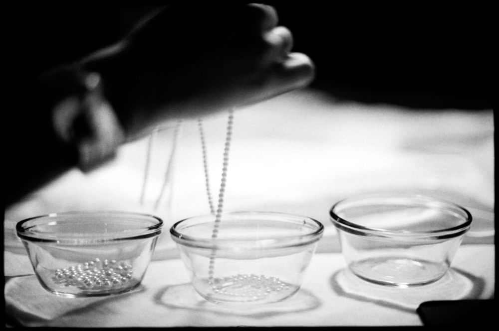 Black and white image of glass bowls and beaded chain on a white surface with hands above.