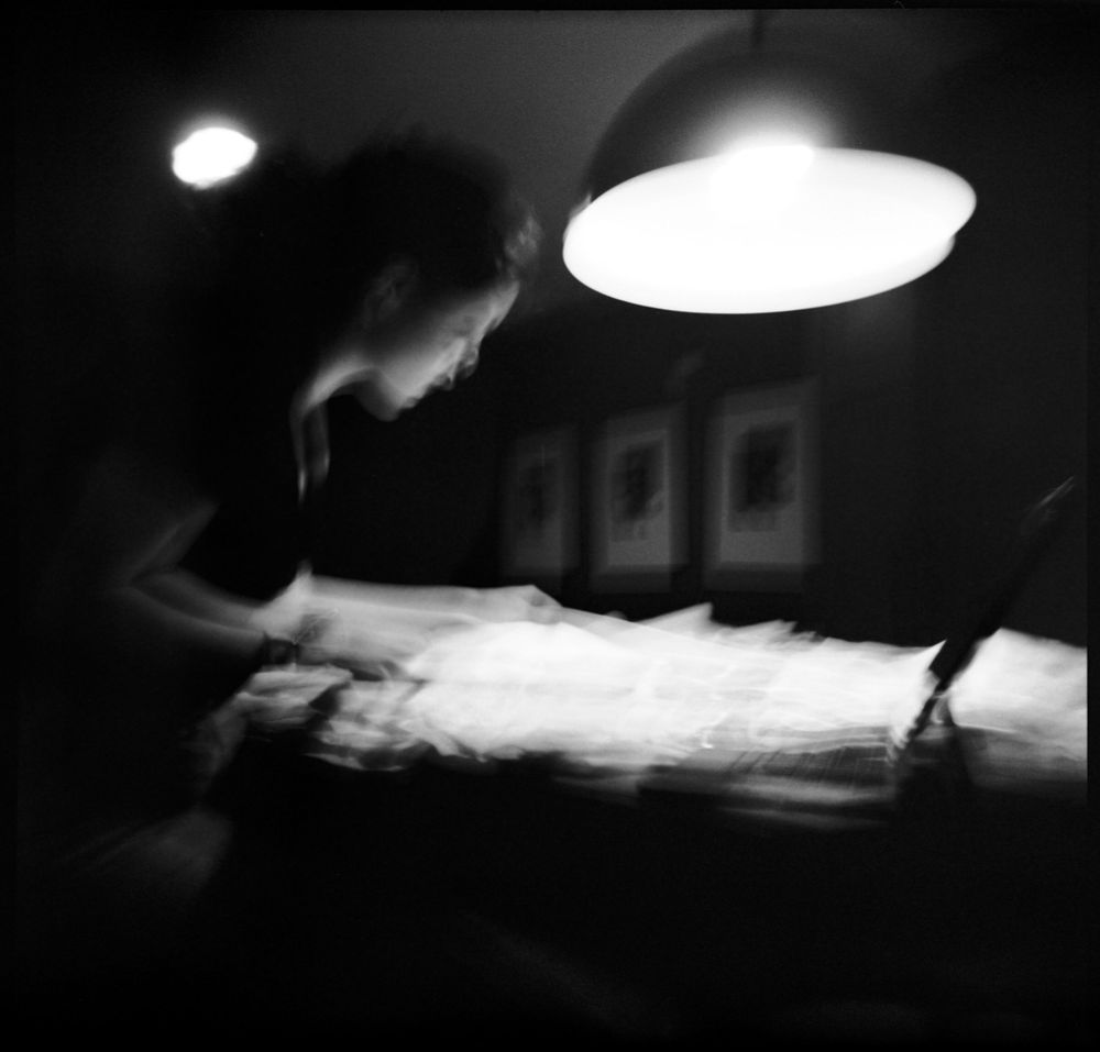 Black and white blurred image of a person working under a lamp over a white fabric table.