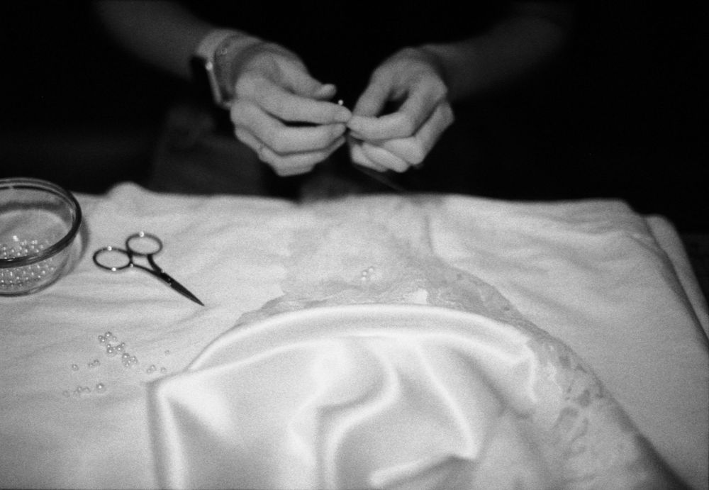 Black and white image of hands threading a needle over white satin fabric with scissors nearby.