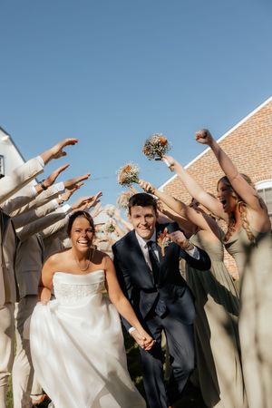 Smiling bride and groom walk through cheering wedding party tunnel outdoors under a clear blue sky.