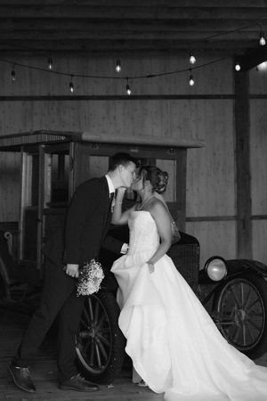 Black and white photo of a bride and groom sharing a kiss in a rustic barn with string lights.