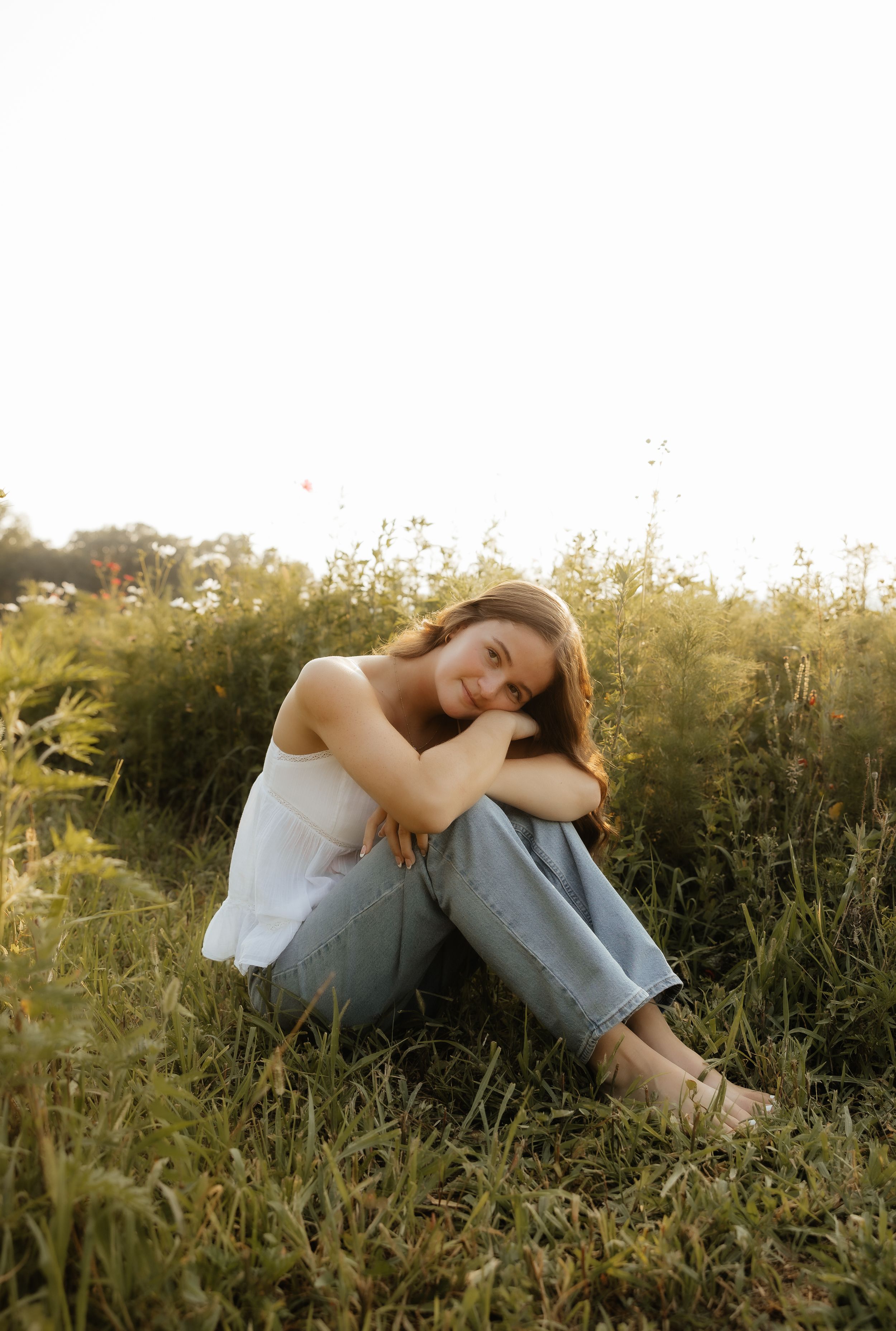 Young woman in white top and jeans sitting in a sunlit meadow, smiling warmly with knees hugged to her chest.