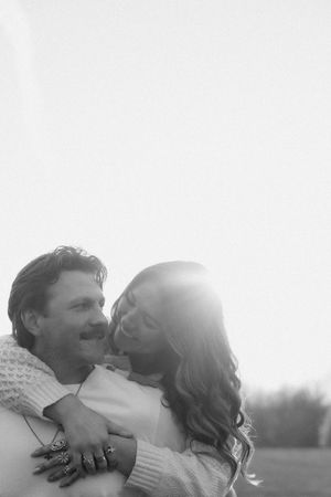 Black and white photo of a laughing couple embracing outdoors with soft backlit sunlight glowing behind them.