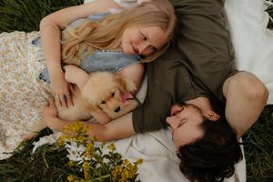 Couple lying on grass with golden puppy and wildflowers in romantic outdoor portrait session.
