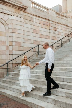 A smiling bride in a white tiered dress holds hands with her groom on stone steps outside a classic building.