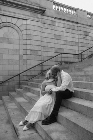 Black and white photo of a couple sharing a romantic kiss while sitting on stone steps outside a classic building.