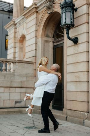 A groom lifts his bride in a white dress outside a grand stone building, sharing a joyful romantic moment.