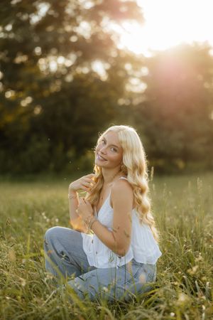 Woman in white top sits in sunlit meadow during golden hour while posing thoughtfully among tall grass.