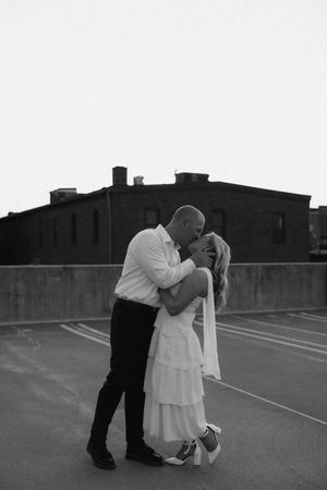 Black and white photo of a couple sharing a romantic kiss in a parking lot, bride in white dress, groom in dark suit.