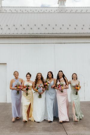 Bride and bridesmaids in pastel gowns holding colorful bouquets pose in front of a white barn.
