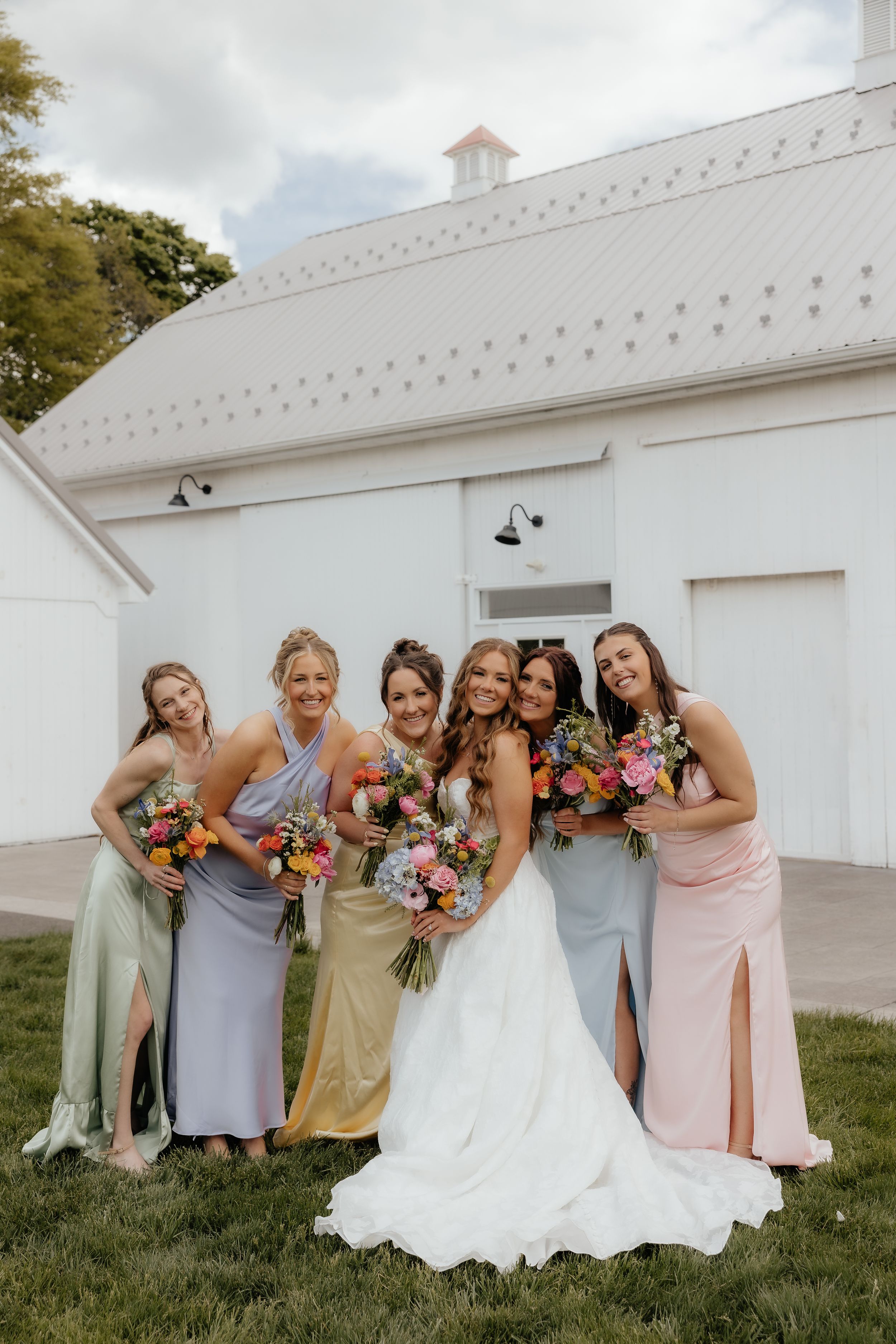 Bride in white gown poses with five bridesmaids in colorful satin dresses holding vibrant floral bouquets outdoors.
