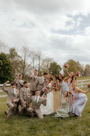 Joyful wedding party celebrating outdoors in pastel attire, bride in white gown holding colorful bouquet on green lawn.