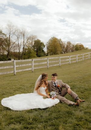 Bride and groom sitting together on green grass field with white fence and trees in background on wedding day.