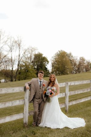 A bride and groom pose by a white wooden fence on a green farm, she holds a colorful bouquet in her flowing white gown.