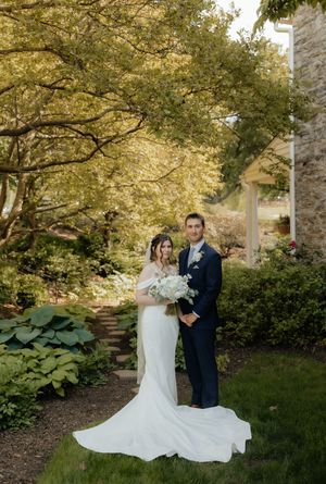 Bride and groom posing in a lush garden setting, bride holding white bouquet in elegant fitted wedding gown.