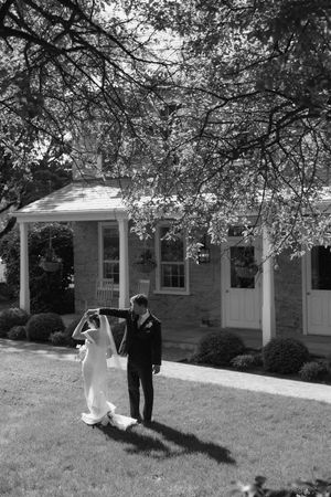 Black and white photo of a bride and groom dancing on a lawn in front of a charming brick house on their wedding day.
