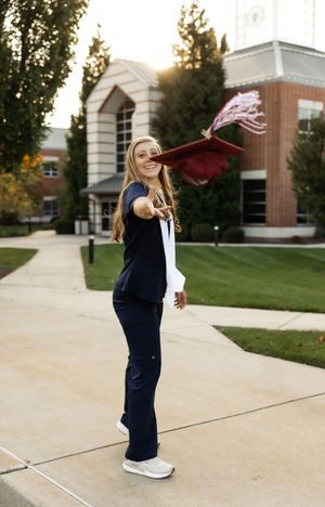 A graduate in navy scrubs tosses their graduation cap outside a brick building.