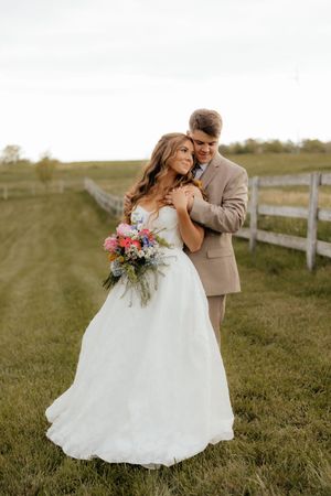Bride and groom embracing in a green field by a wooden fence, bride holding colorful bouquet in white gown.