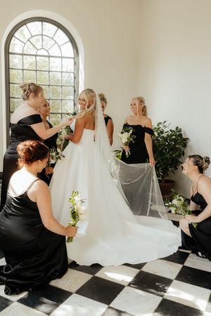 Bridesmaids in black dresses help adjust a flowing white wedding gown and veil in a sunlit room with checkered floors.