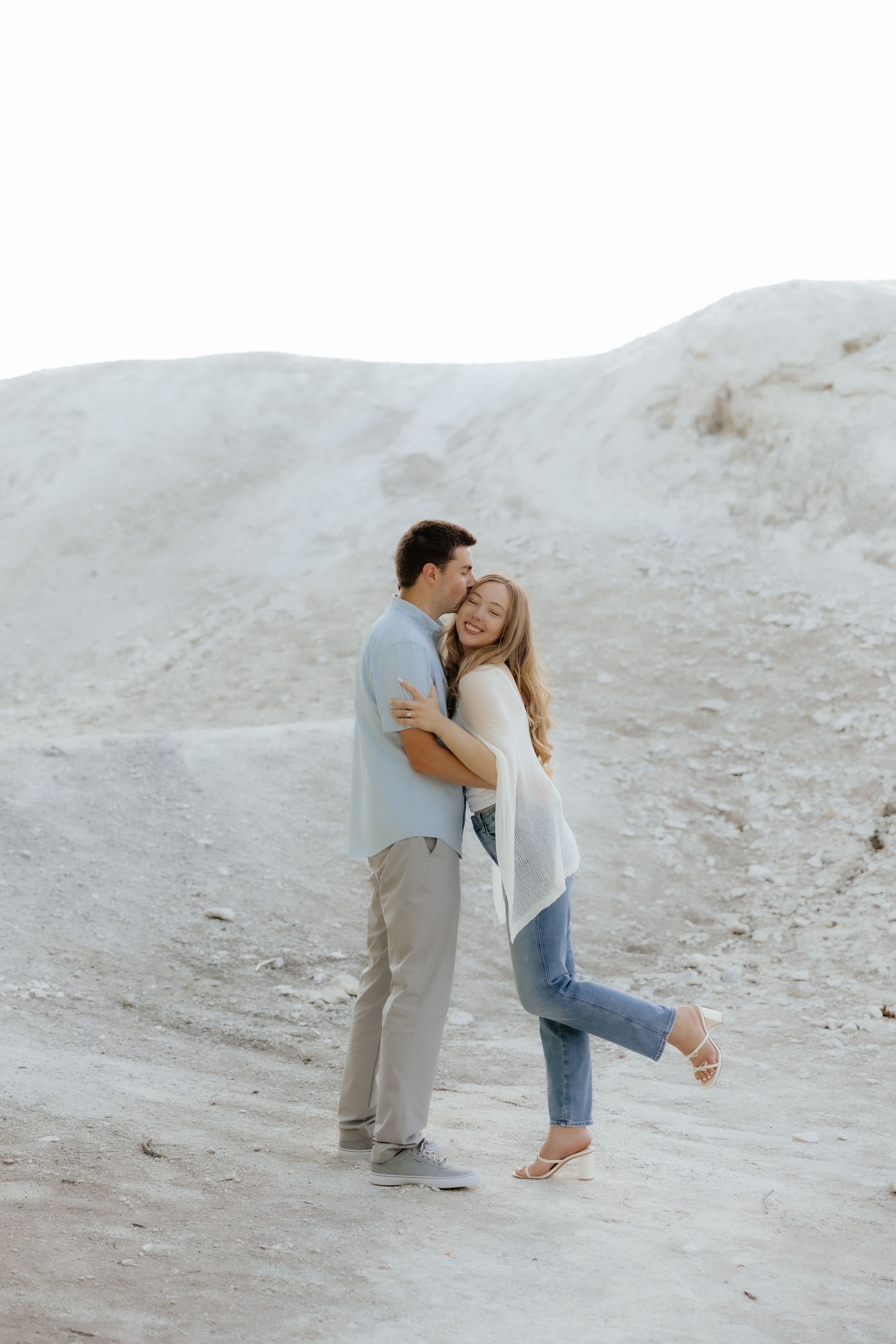 A couple embraces and shares a kiss on sandy dunes, the woman smiling joyfully in jeans and white cardigan.
