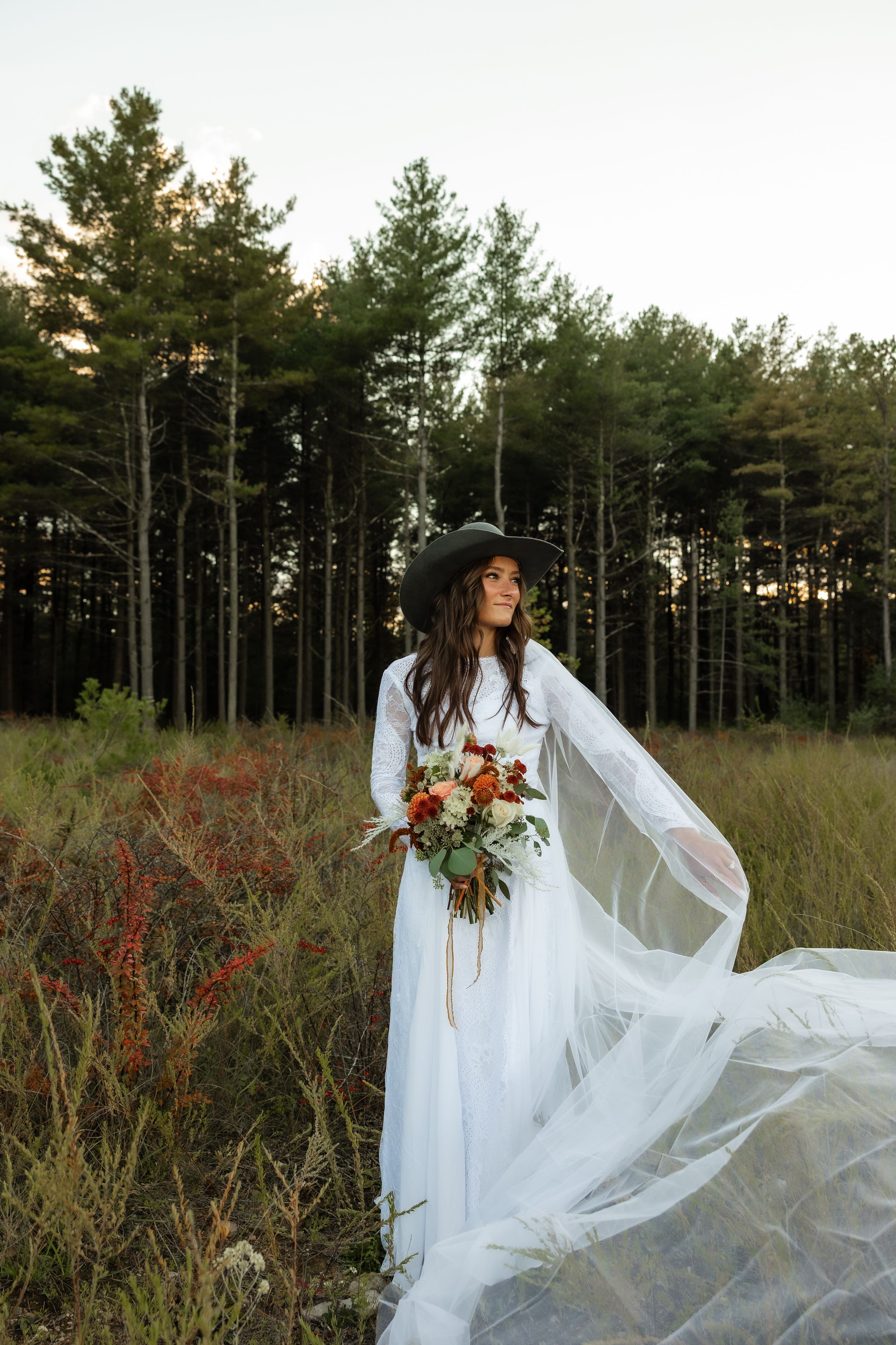 A bride in a flowing white dress and hat stands in a field with pine trees at sunset.