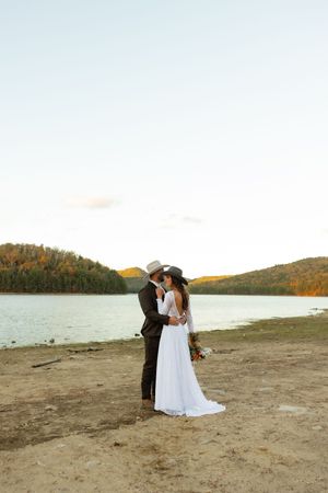 A newlywed couple in western attire shares an intimate moment by a lake at sunset.
