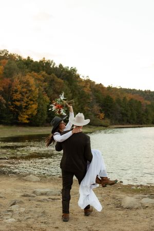 A couple embraces on a lakeside beach surrounded by autumn foliage.