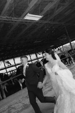 Black and white photo of a bride and groom holding hands, laughing as they walk through a rustic barn wedding venue.