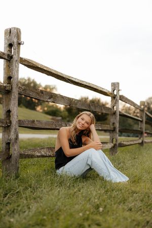 A person in a black top and light jeans sits casually by a rustic wooden fence in a grassy field at sunset.